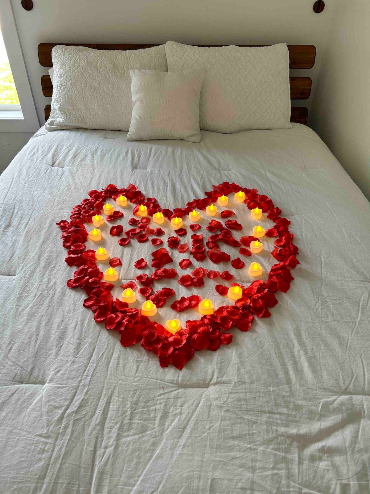 A heart shape formed by red rose petals is displayed on a white bedspread. Small LED candles are arranged within the petals, creating a soft glow. Two decorative cushions placed against the headboard enhance the inviting look of the bed.