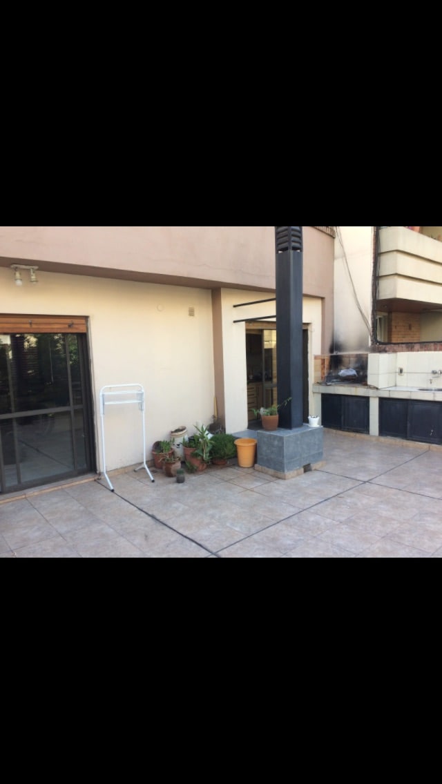 A spacious terrace is shown, featuring tiled flooring and a stone planter with greenery. An outdoor grill is positioned near the corner, and a small drying rack stands nearby. Large glass doors provide access to the interior living areas.