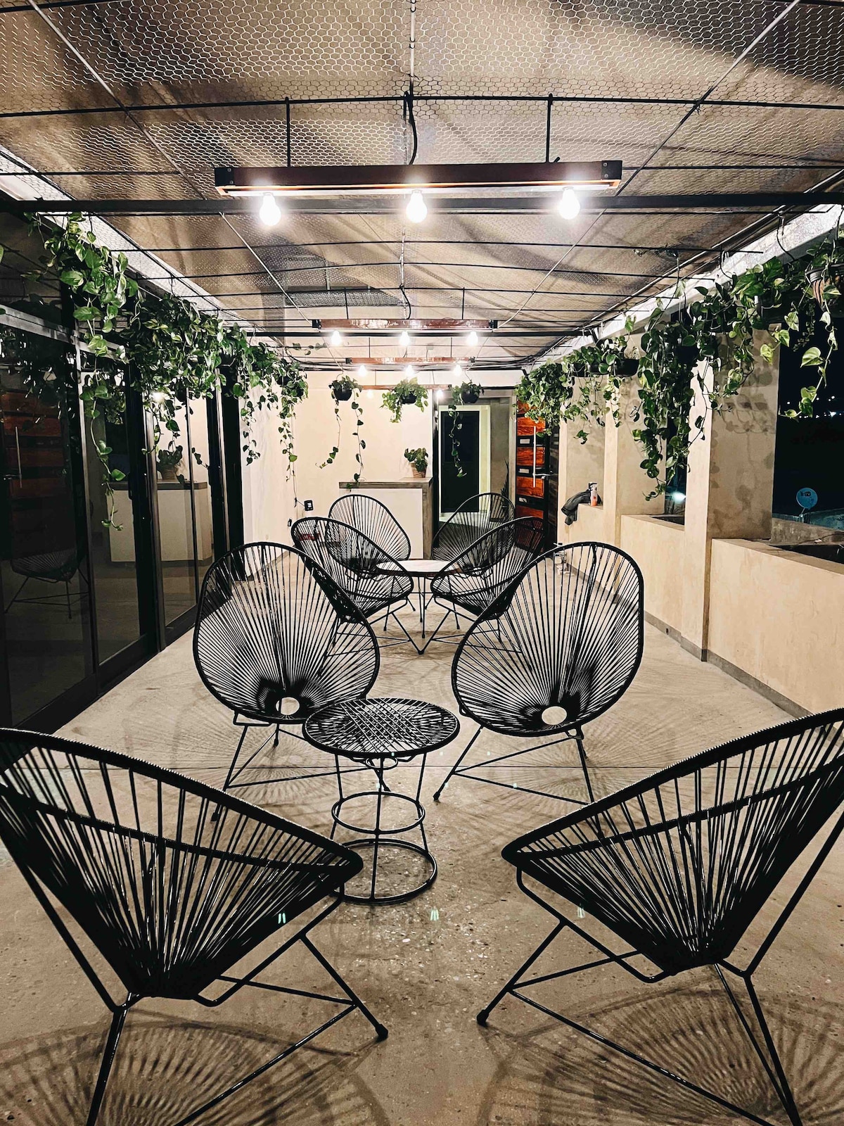 A covered terrace is furnished with several black wire chairs arranged around a small table. Green plants hang from the ceiling, and large glass doors allow natural light to fill the space.
