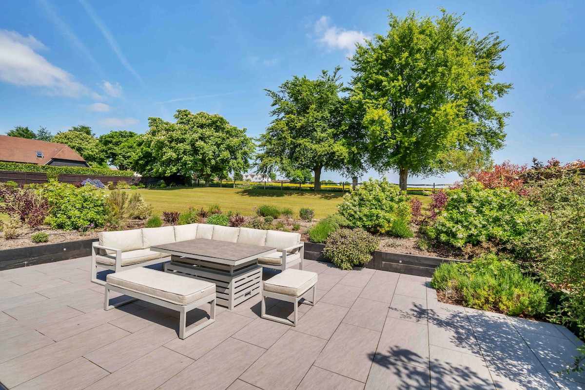A spacious outdoor seating area is shown on a stone patio, featuring a large rectangular table surrounded by several light-colored benches. Lush greenery and flowering plants create a natural border, with large trees providing shade in the background under a clear blue sky.