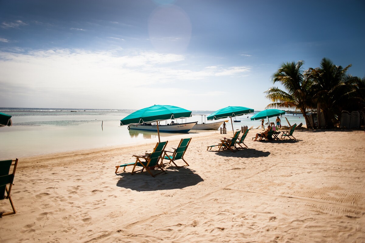 A sandy beach area features several wooden lounge chairs, positioned under teal umbrellas. Gentle waves lap at the shore, with small boats anchored in the water. Lush palm trees provide shade in the background, enhancing the tranquil coastal environment.