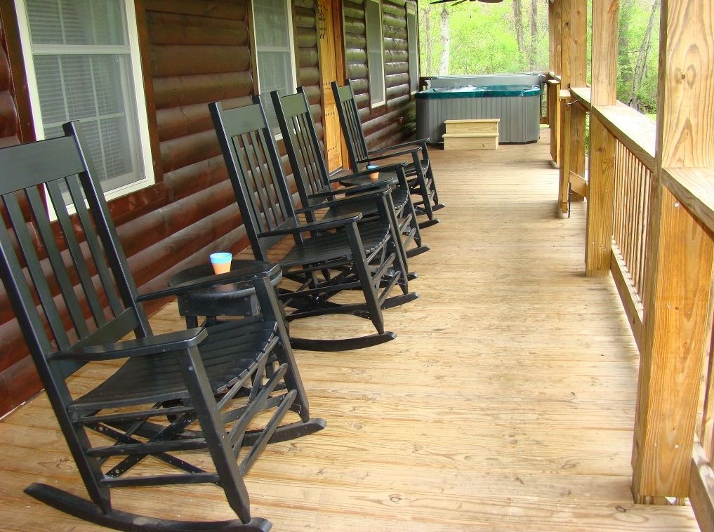 A spacious front porch features multiple black rocking chairs arranged along the railing. A hot tub is visible in the background, surrounded by the natural landscape. The wooden floor and log cabin walls create a rustic yet inviting ambiance.