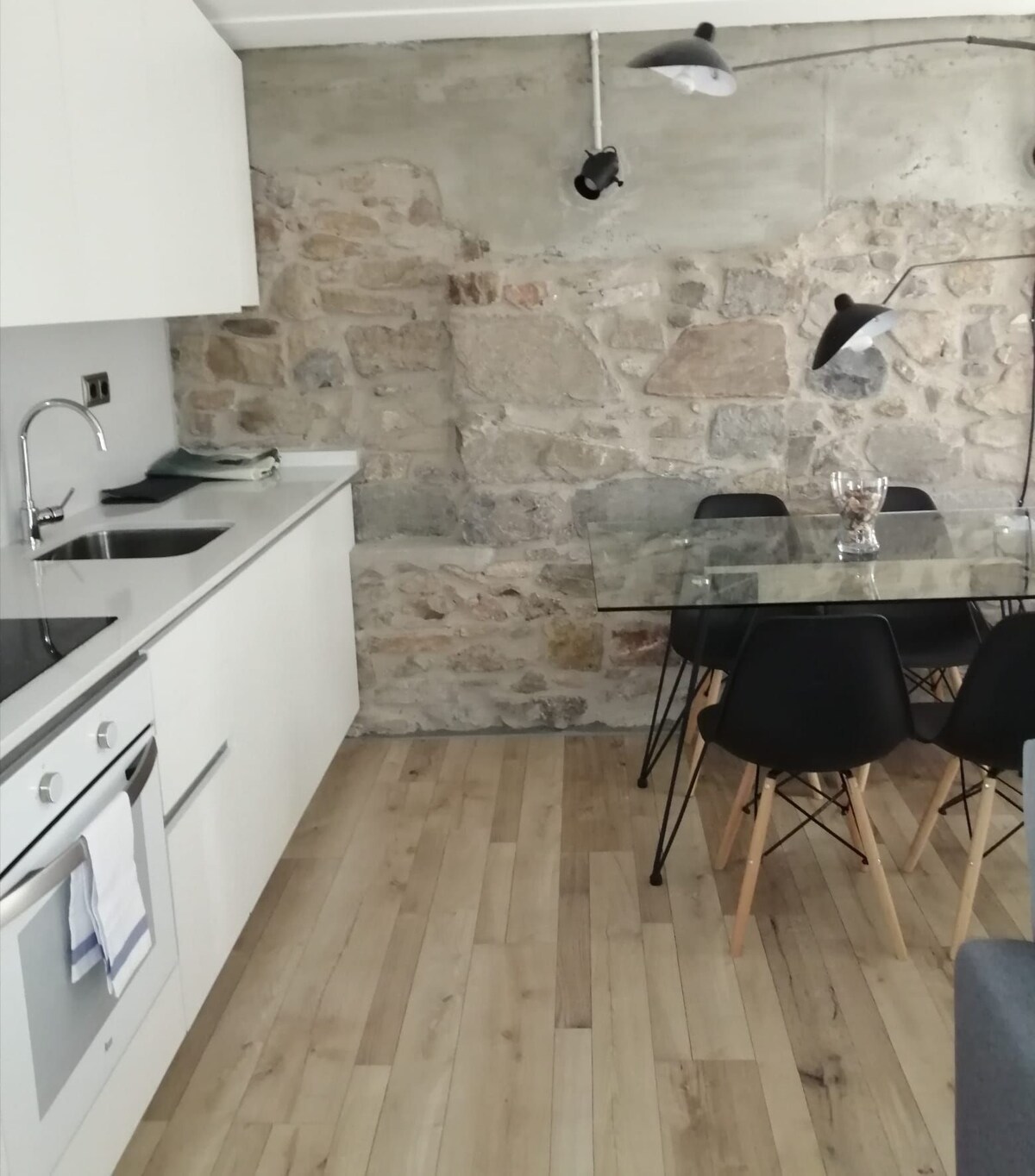 A modern kitchen area features sleek white cabinetry paired with a glass-topped dining table and black chairs. Exposed stone walls add texture, while wood flooring contributes to a warm aesthetic. Ambient lighting is provided by wall-mounted fixtures, enhancing the inviting atmosphere.