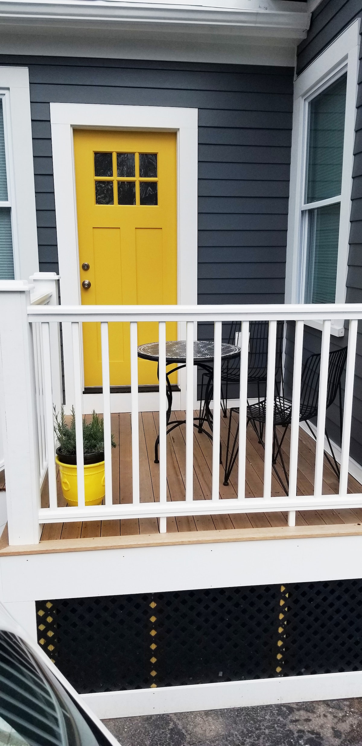 An inviting porch area features a vibrant yellow door that stands out against gray siding. Two black chairs and a small table offer a cozy spot for outdoor relaxation. A potted plant adds a touch of greenery, complementing the bright entrance.
