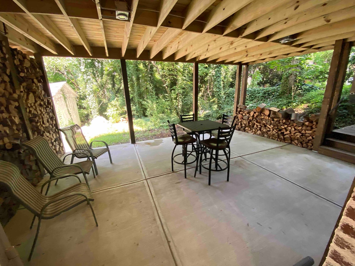A shaded outdoor area features a metal table surrounded by four black chairs. Stacked firewood lines one side, while greenery surrounds the space, creating a natural ambiance. The concrete floor provides a clean and functional surface.