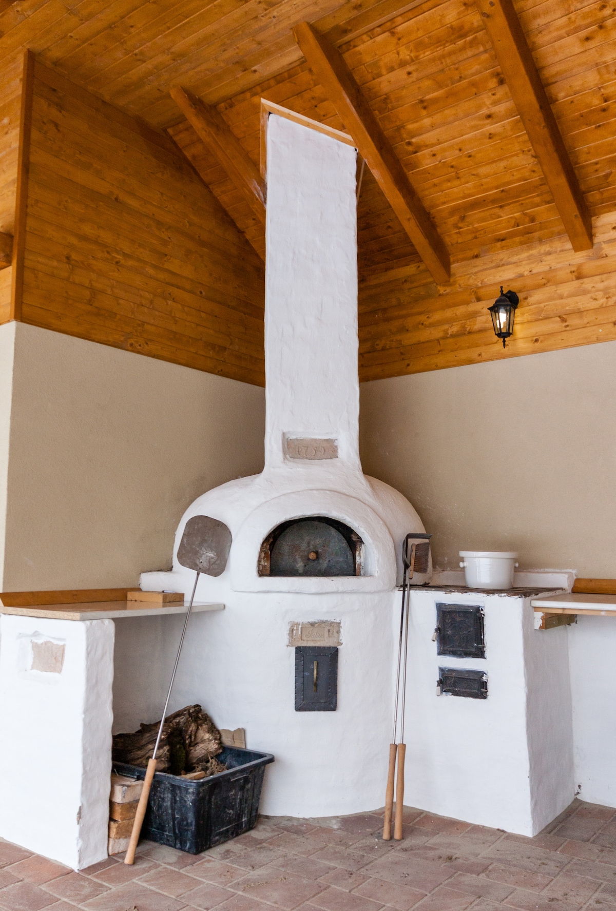An outdoor oven features a white stucco exterior with a domed shape, supported by a wooden ceiling with exposed beams. Adjacent workspace includes wooden counters, a wood storage container, and cooking tools, offering a practical area for outdoor cooking and gatherings.