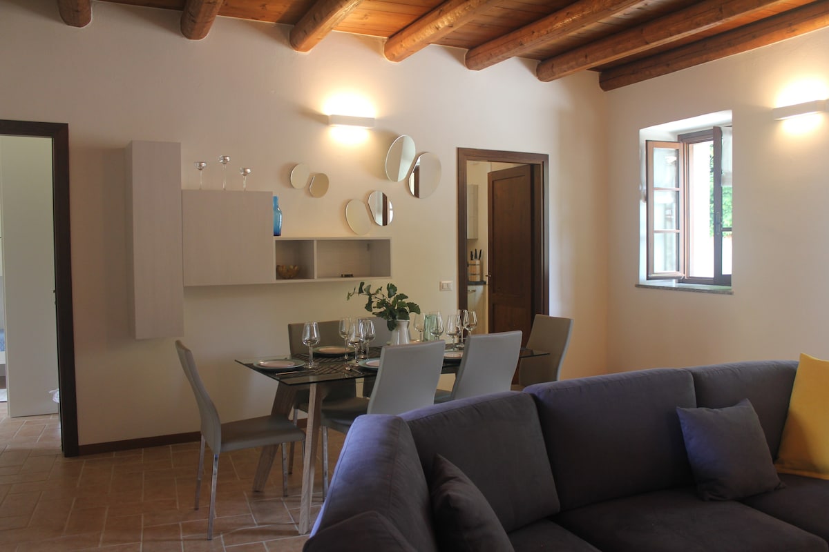 A bright dining area features a modern glass table set for four, complemented by simple grey chairs. The wall is decorated with round mirrors, and natural light enters through a window, enhancing the inviting atmosphere. A comfortable grey couch is positioned nearby.