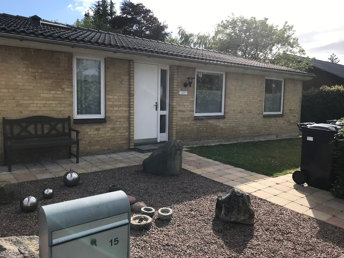The exterior of a brick bungalow is displayed, featuring a welcoming entrance with a door beside two large windows. A stone walkway is visible, bordered by small rocks and greenery. A bench is positioned on the left, adding a functional outdoor seating area.