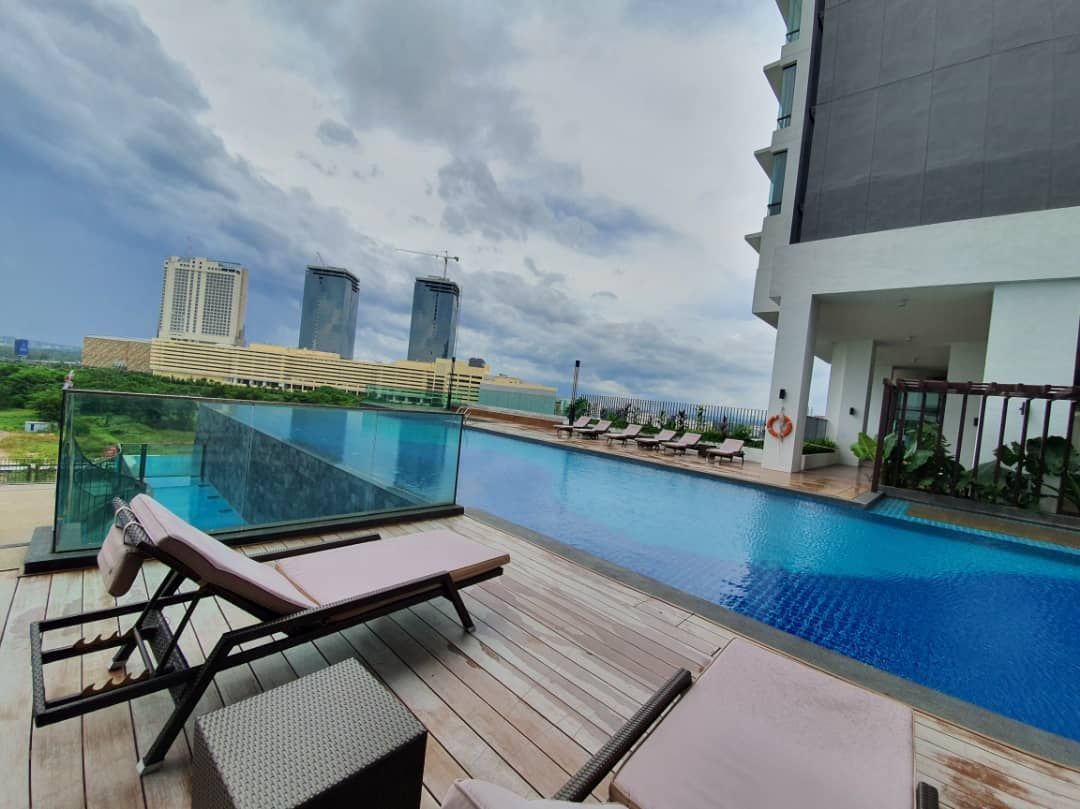 An infinity pool is highlighted, featuring a sleek edge overlooking a scenic city skyline. Comfortable lounge chairs are positioned nearby on a wooden deck, surrounded by greenery. The cloudy sky adds depth to the tranquil atmosphere, enhancing the overall appeal of the space.