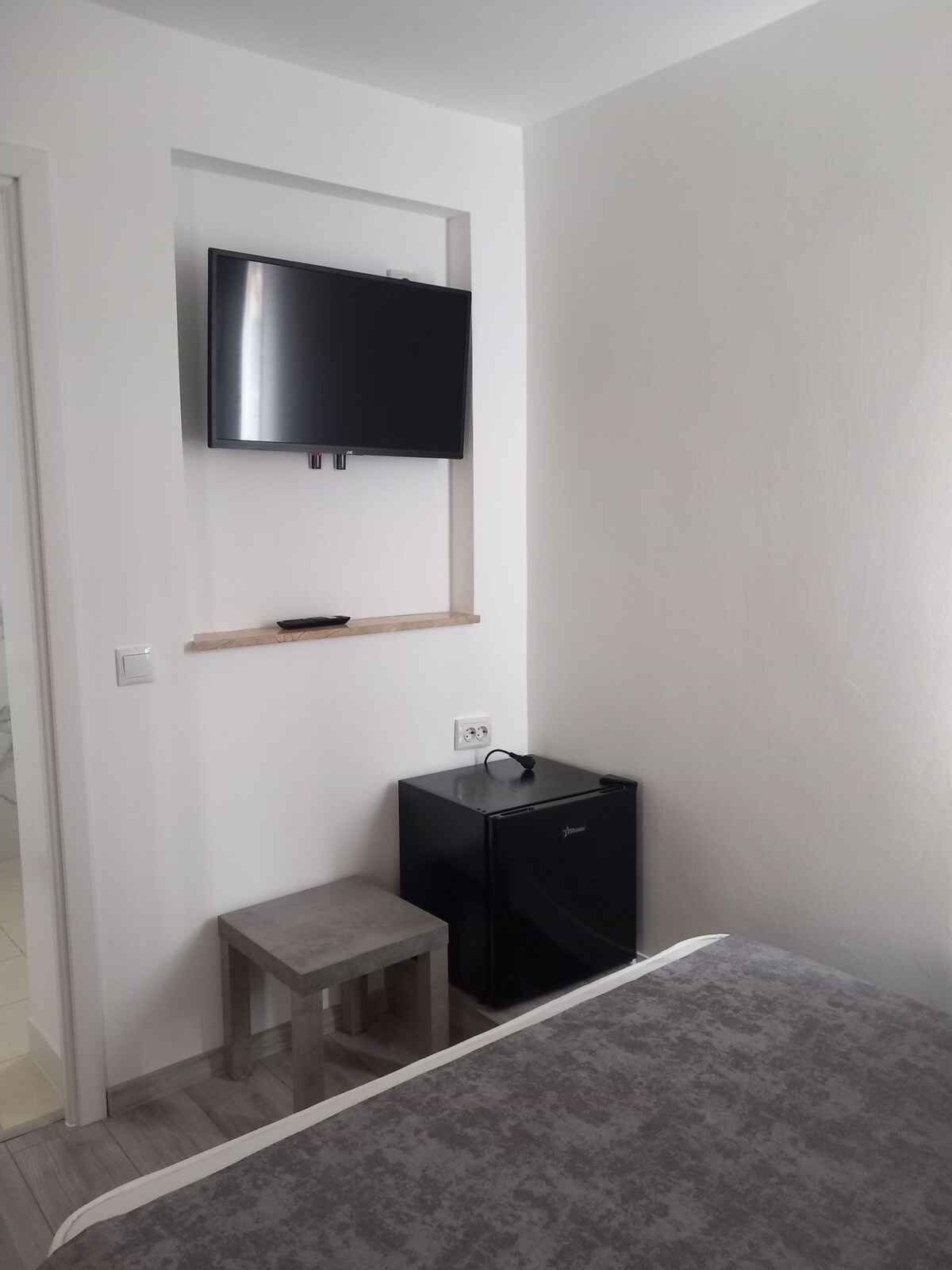 A compact space features a mounted flat-screen TV above a small wooden shelf. A dark mini-fridge rests beside a gray stool, contributing to the room's minimalist design. The light-colored walls enhance the overall brightness of the area.