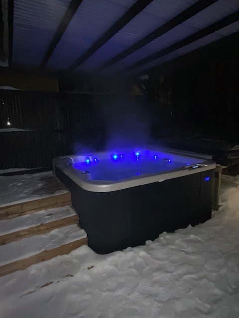 An outdoor hot tub is situated on a wooden deck, surrounded by freshly fallen snow. Soft blue lights illuminate the water, creating a serene ambiance. Steam rises from the tub, suggesting a warm and inviting experience amidst the winter landscape.