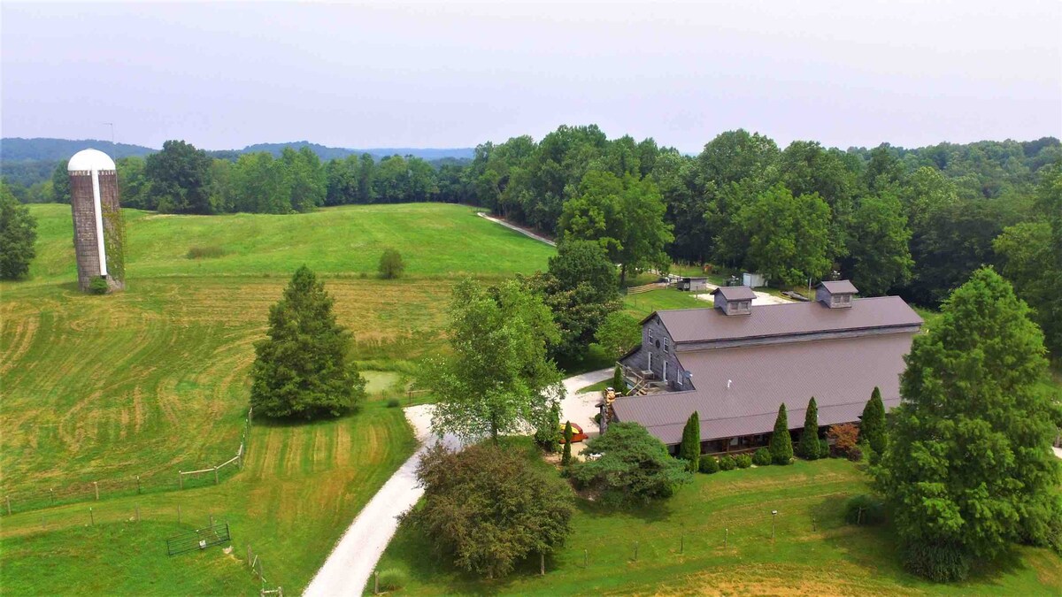 Aerial view of a scenic farm landscape, showcasing a large gray barn and silo amidst lush greenery. A winding driveway leads to the cabin, surrounded by trees and open fields, creating a peaceful rural atmosphere.