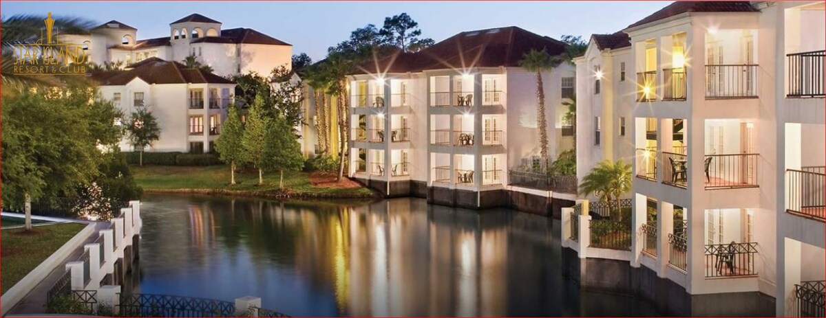 A serene waterfront view showcases a charming resort building with balconies facing a tranquil lake. Soft lighting reflects from the water's surface, and lush greenery surrounds the space, enhancing the natural beauty of the setting.