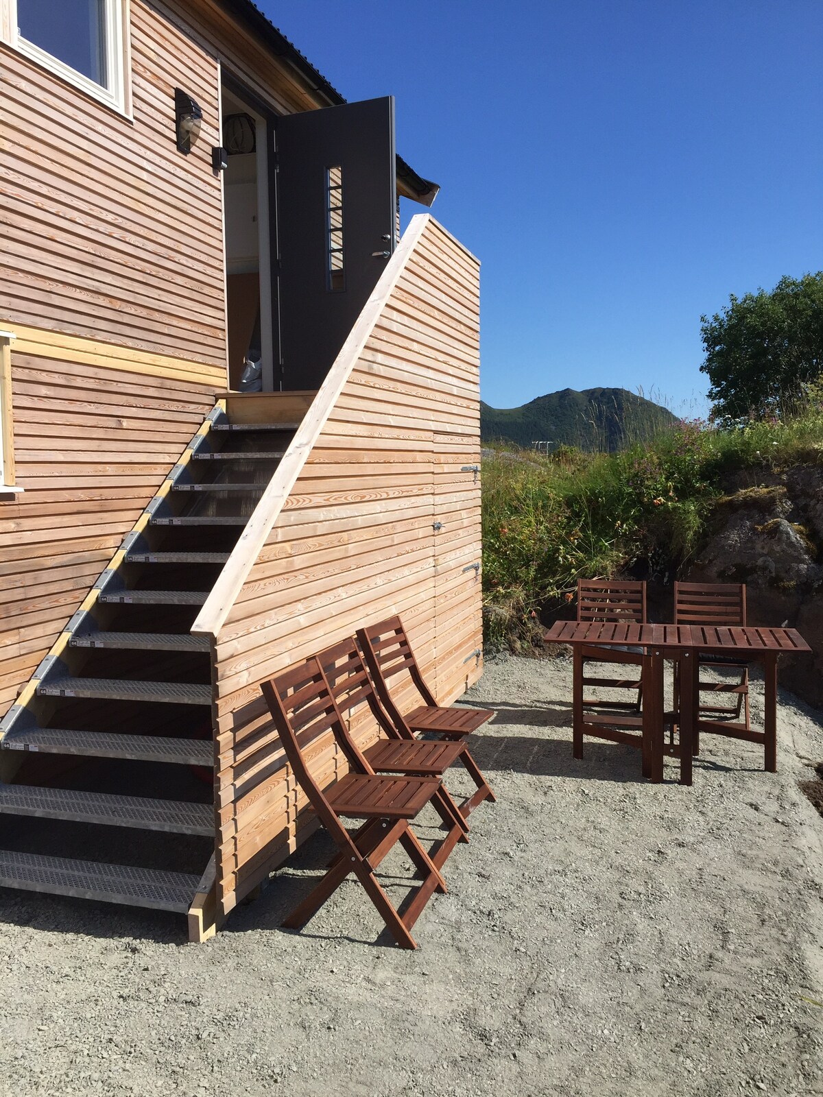 An exterior view showcases a modern wooden structure with stairs leading to a door. A table and four folding chairs are arranged on the gravel area, providing an inviting space for outdoor gatherings. Lush greenery and distant hills are visible in the background.