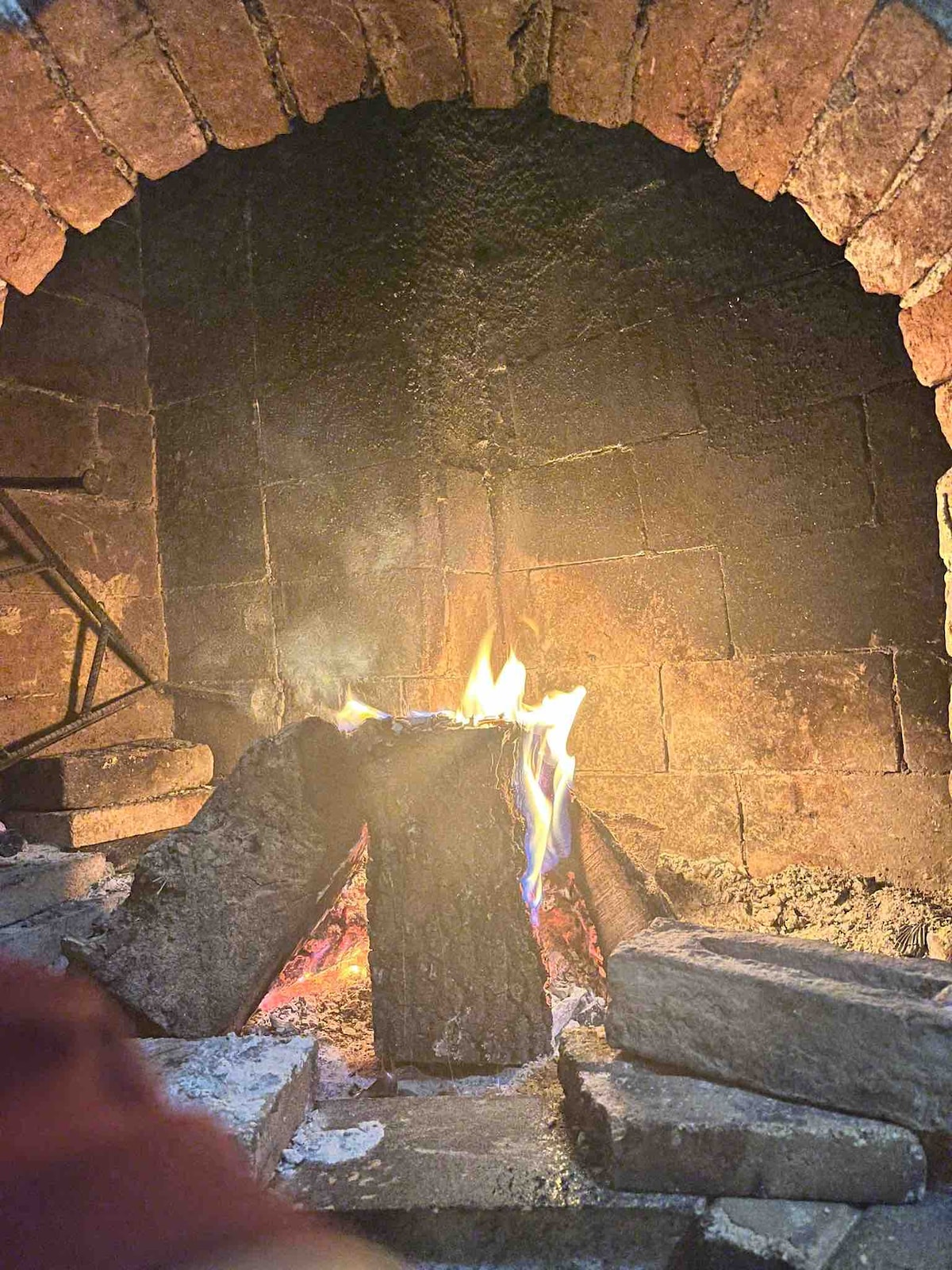 A rustic stone fireplace is shown, with flames flickering against the backdrop of dark, textured bricks. Logs are stacked in a triangular formation at the center, surrounded by scattered pieces of stone. Soft smoke is visible rising from the fire.