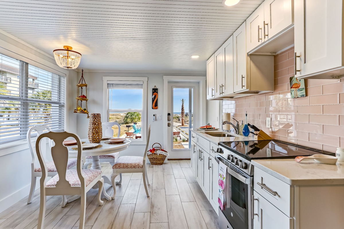 A bright and airy kitchen is equipped with modern stainless steel appliances and quartz countertops. A small dining table with two chairs is positioned near a window, showcasing views of the outdoor space. Natural light fills the room, enhancing the inviting atmosphere.