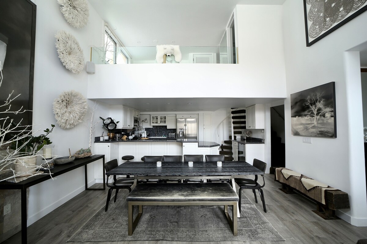 An open-concept dining area features a long wooden table accompanied by dark chairs, positioned under high ceilings. Light walls and large windows create a bright ambience. The adjoining kitchen and living space are visible in the background, enhancing the spacious feel of the room.