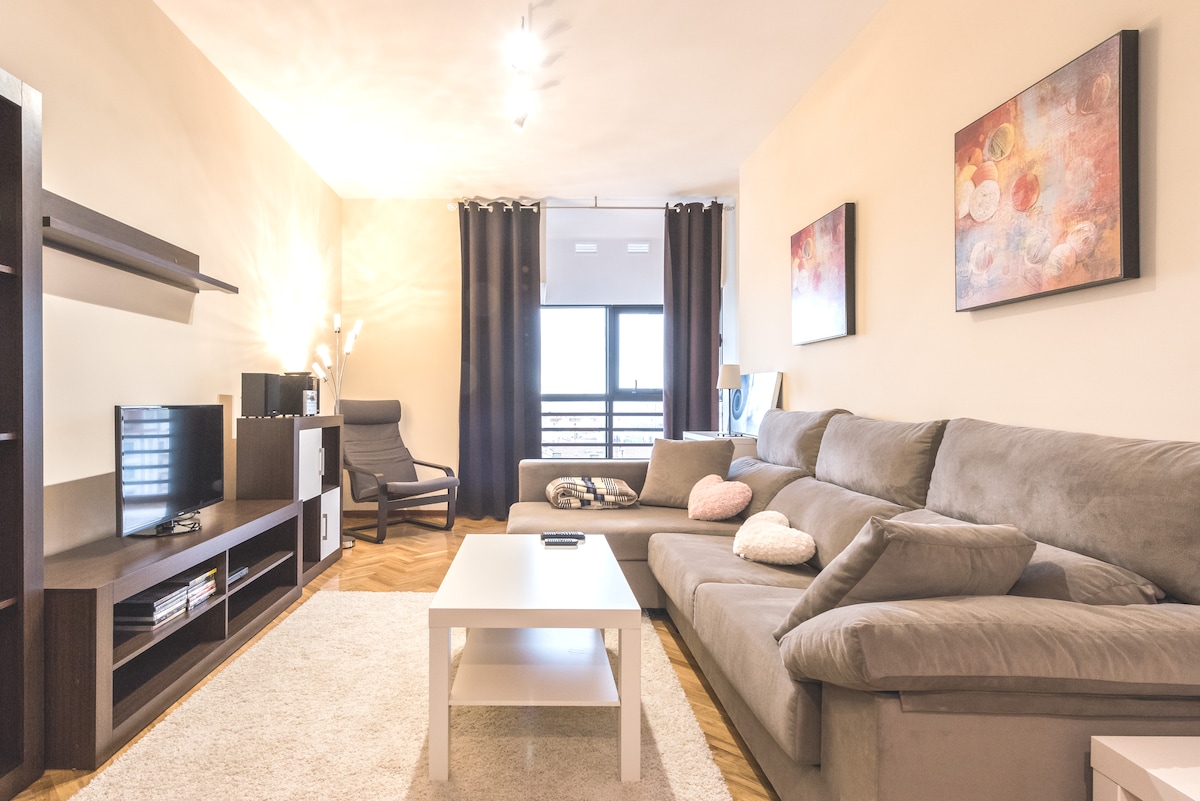 A spacious living area is furnished with a large, comfortable sectional sofa and a light-colored coffee table. A soft rug covers the wooden floor. A TV unit is positioned against the wall, complemented by artistic wall decor and large windows dressed with dark curtains.