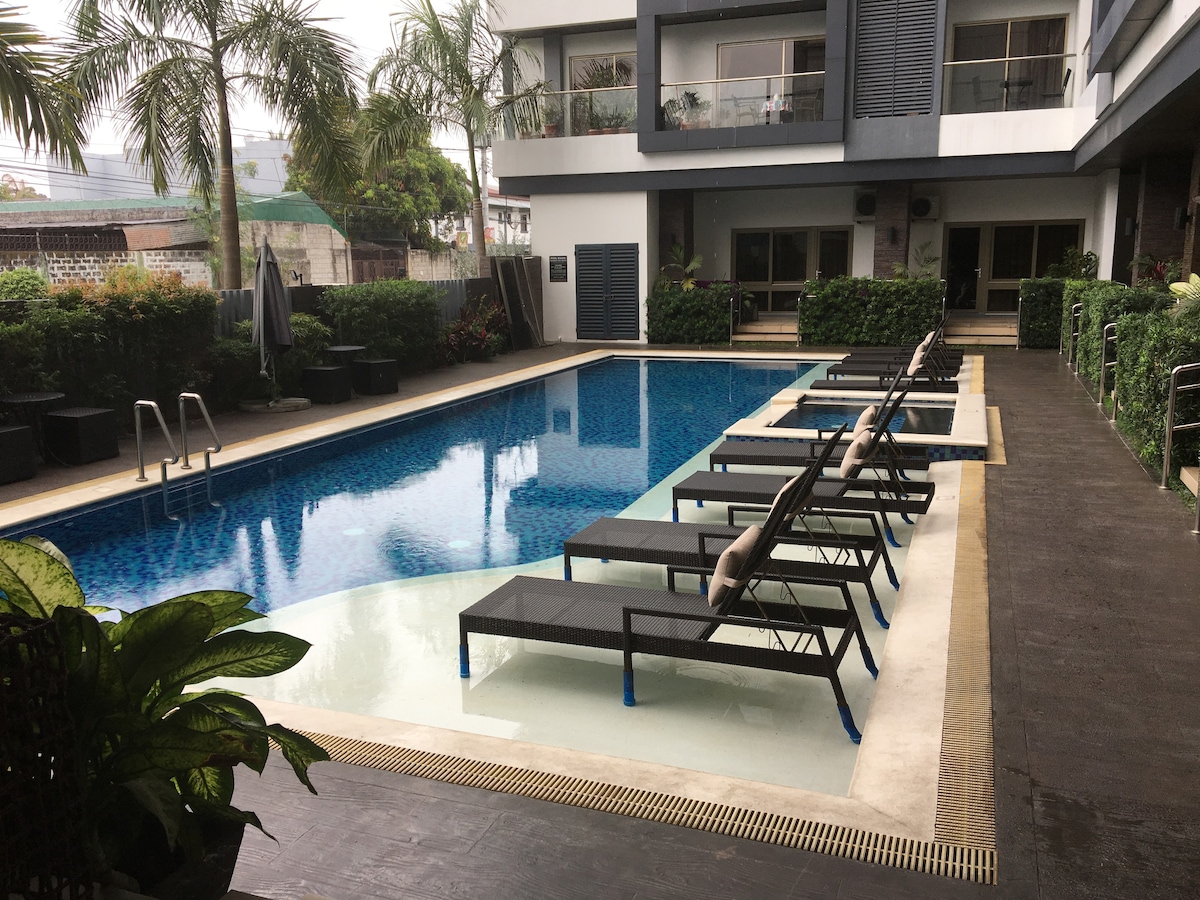 A swimming pool area features a series of lounge chairs positioned alongside the water. Lush greenery surrounds the space, with palm trees adding to the atmosphere. A shaded area with plants is visible in the background, along with comfortable seating options.