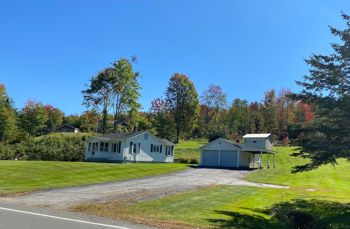 A charming bungalow is surrounded by expansive green lawn, featuring a paved driveway leading to a double garage. Trees with colorful autumn foliage create a picturesque backdrop against the clear blue sky, enhancing the inviting rural setting.