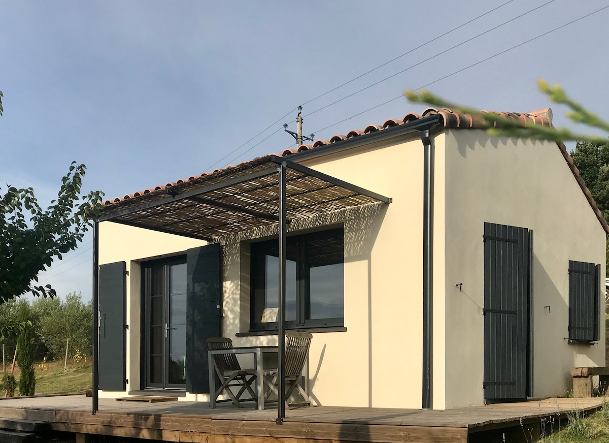 A charming single-story home features a covered terrace with a table and chairs. Large windows allow natural light to enter, and dark shutters can be seen closed on either side. The surrounding landscape includes trees and open space.