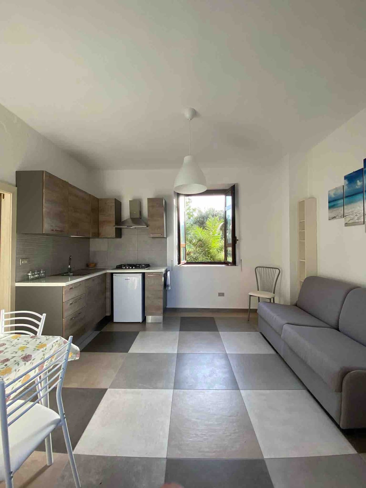 The image shows a modern kitchen and living area featuring a grey sofa and a dining table with white chairs. A window allows natural light to enter, revealing views of greenery outside. The floor displays a checkered pattern in neutral tones.
