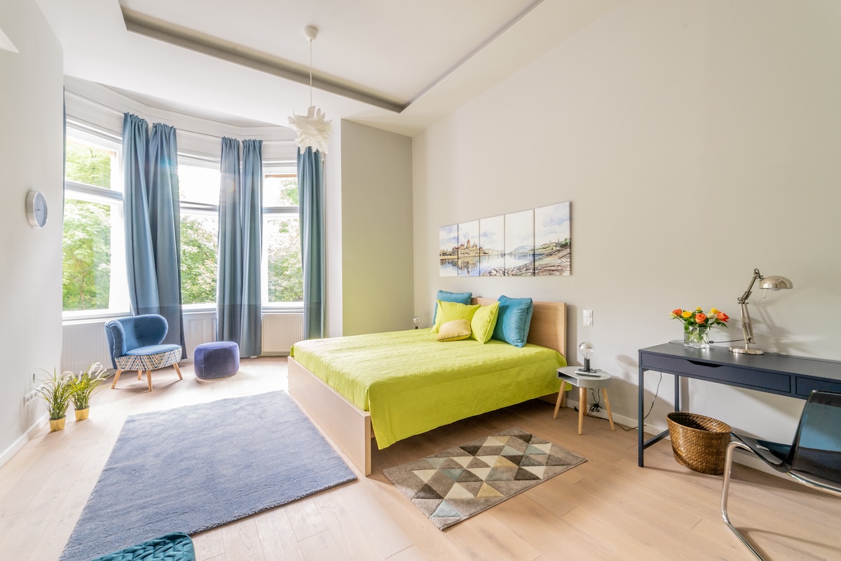 A spacious bedroom is highlighted with large windows dressed in light blue curtains. A vibrant green bedspread covers a comfortable bed, accompanied by decorative pillows. A cozy seating area features a blue armchair and a small table. A desk with a lamp and a basket add functional touches.
