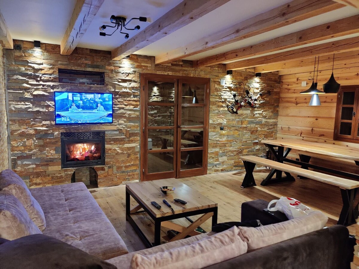 A rustic living area features a combination of stone and wooden walls, enhancing the natural ambiance. A flat-screen television is mounted above a modern fireplace. Stylish pendant lights hang above a long wooden dining table, which is complemented by a comfortable sofa arrangement.