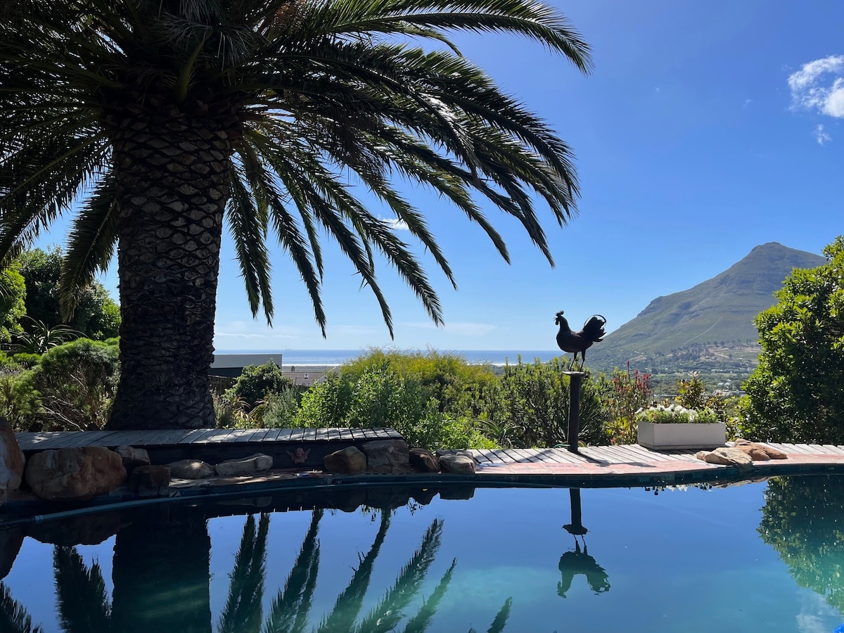 A tranquil pool is framed by lush greenery and a towering palm tree, with a sculpture of a rooster visible in the foreground. The view showcases the expansive landscape and mountains against a clear blue sky.