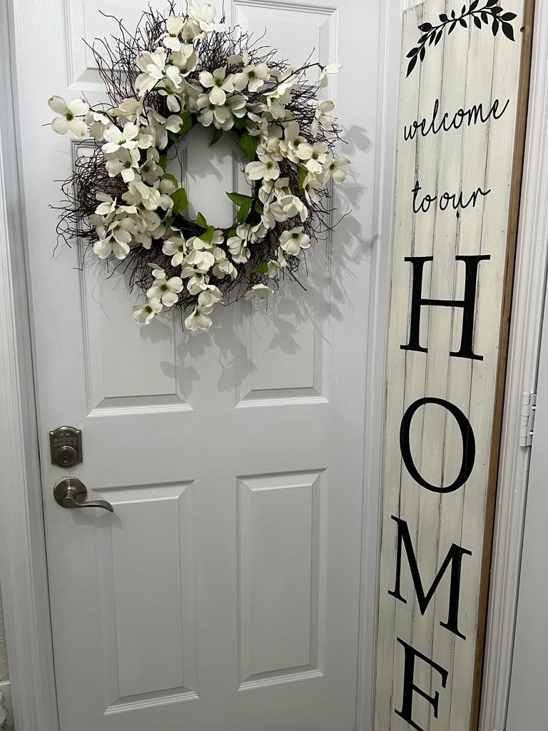 A white door is adorned with a decorative wreath featuring white flowers and greenery. Adjacent to the door, a wooden sign with a rustic finish displays the word 'HOME' in large, bold letters, creating a welcoming entryway.
