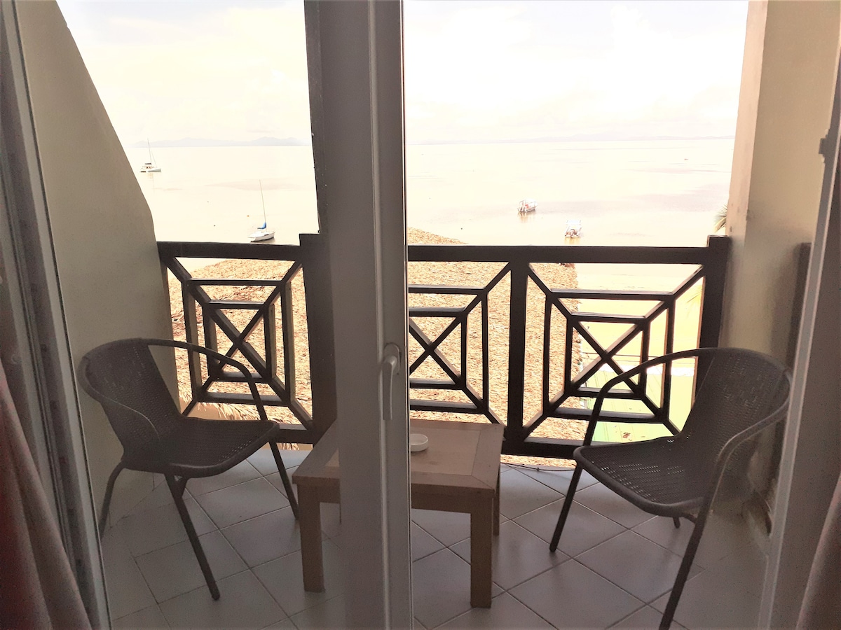 A balcony is visible, featuring two black woven chairs and a small wooden table. The railing is decorated with geometric patterns. Beyond the balcony, a serene view of the ocean and boats can be seen, emphasizing a tranquil outdoor space.