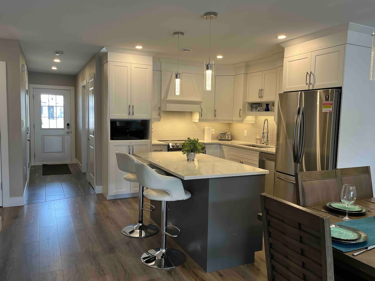 The kitchen features modern cabinetry and stainless steel appliances, complemented by a spacious island with seating for four. Natural light floods the room through a window, highlighting the sleek countertops and a cozy dining area nearby.