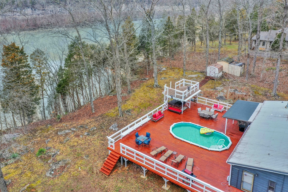 A spacious outdoor deck features a circular pool surrounded by lounge chairs and seating areas. The lush, wooded surroundings provide a natural backdrop, and the nearby lake is visible, emphasizing the tranquility of the lakeside setting.