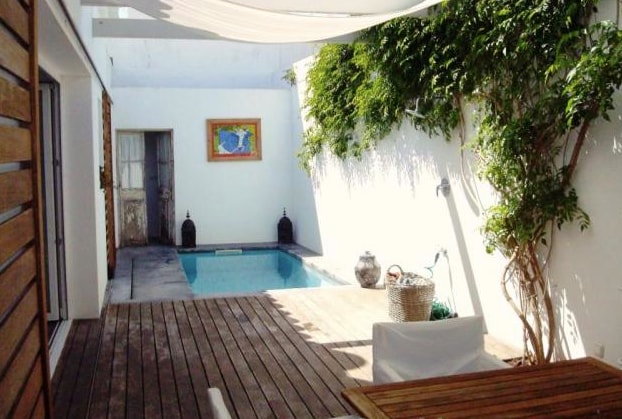 A serene outdoor space features a private pool surrounded by wooden decking. Lush greenery climbs the white walls, while sunlight filters through a canopy. A decorative wall art piece and a few ornamental objects are visible, contributing to a tranquil atmosphere.