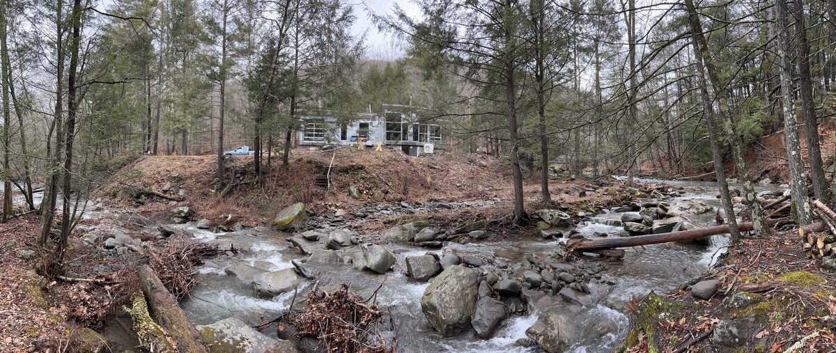 A serene river flows through a wooded area, bordered by smooth stones and fallen branches. In the background, a modern home with large windows is nestled among the trees, offering a peaceful retreat by the water.