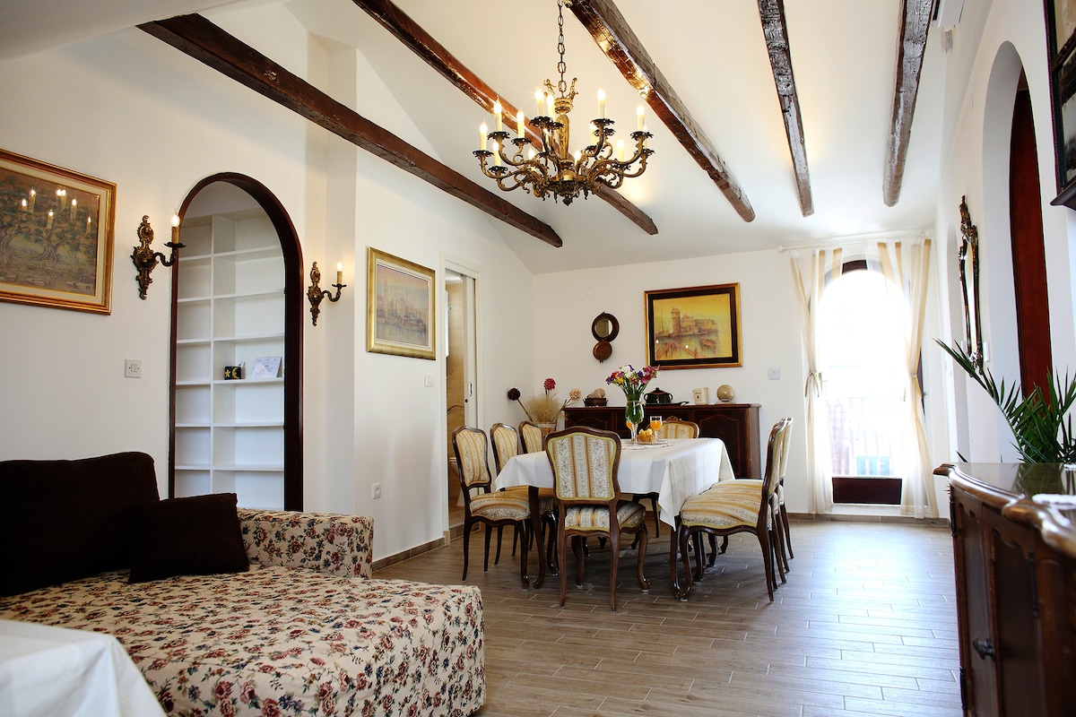 A spacious living area is displayed, featuring exposed wooden beams across the ceiling. A dining table with ornate chairs occupies the center, while a cozy seating area with a floral sofa is visible. Natural light is introduced through large windows, enhancing the inviting atmosphere.