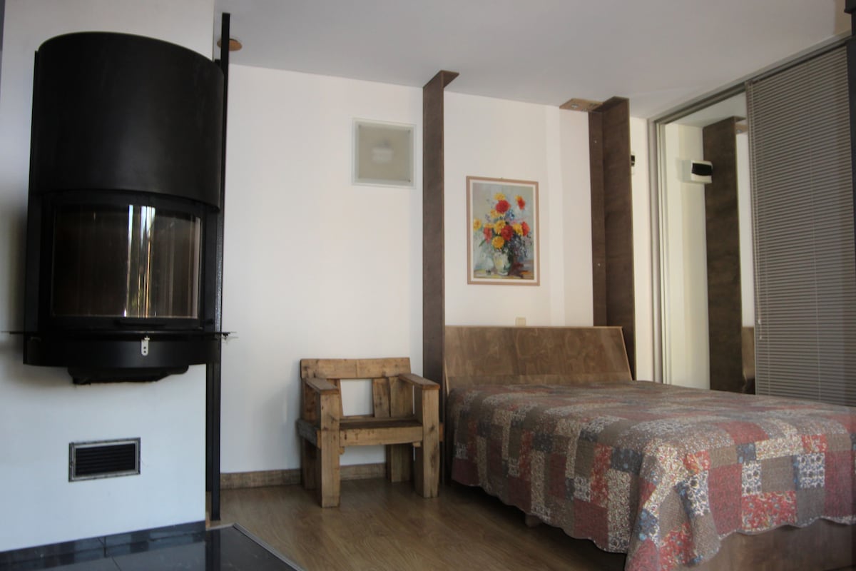 A modern studio apartment features a designer wall-mounted fireplace with a glass door, situated adjacent to a bed draped with a colorful quilt. Natural light filters in through a large, partially glass wall, enhancing the room's aesthetic.