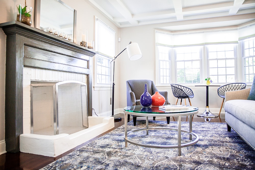 The living room features a decorative fireplace, accented with a round glass coffee table. Comfortable seating includes a gray armchair and a sectional sofa. Natural light streams through multiple large windows, illuminating the space adorned with patterned rugs and simple decor elements.