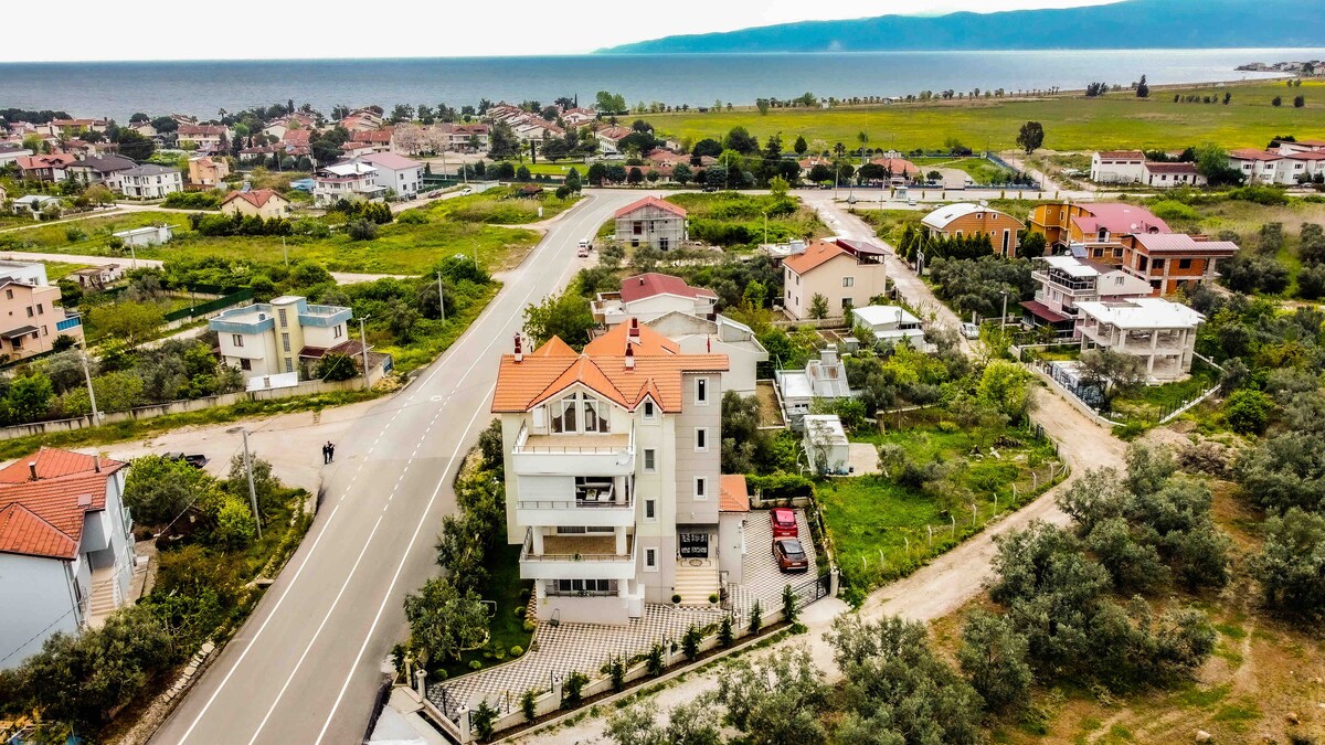 An aerial view captures the spacious property situated between the mountain and the sea, showcasing its modern structure with a red-tiled roof. Surrounding greenery and residential areas are visible, along with the distant coastline and calm blue waters.