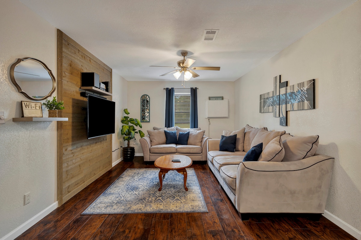 A spacious living area features a comfortable sectional sofa with decorative pillows, positioned near a coffee table. A flat-screen TV is mounted on a wooden accent wall. Natural light enters through windows, adorned with dark curtains, and a potted plant adds a touch of greenery.