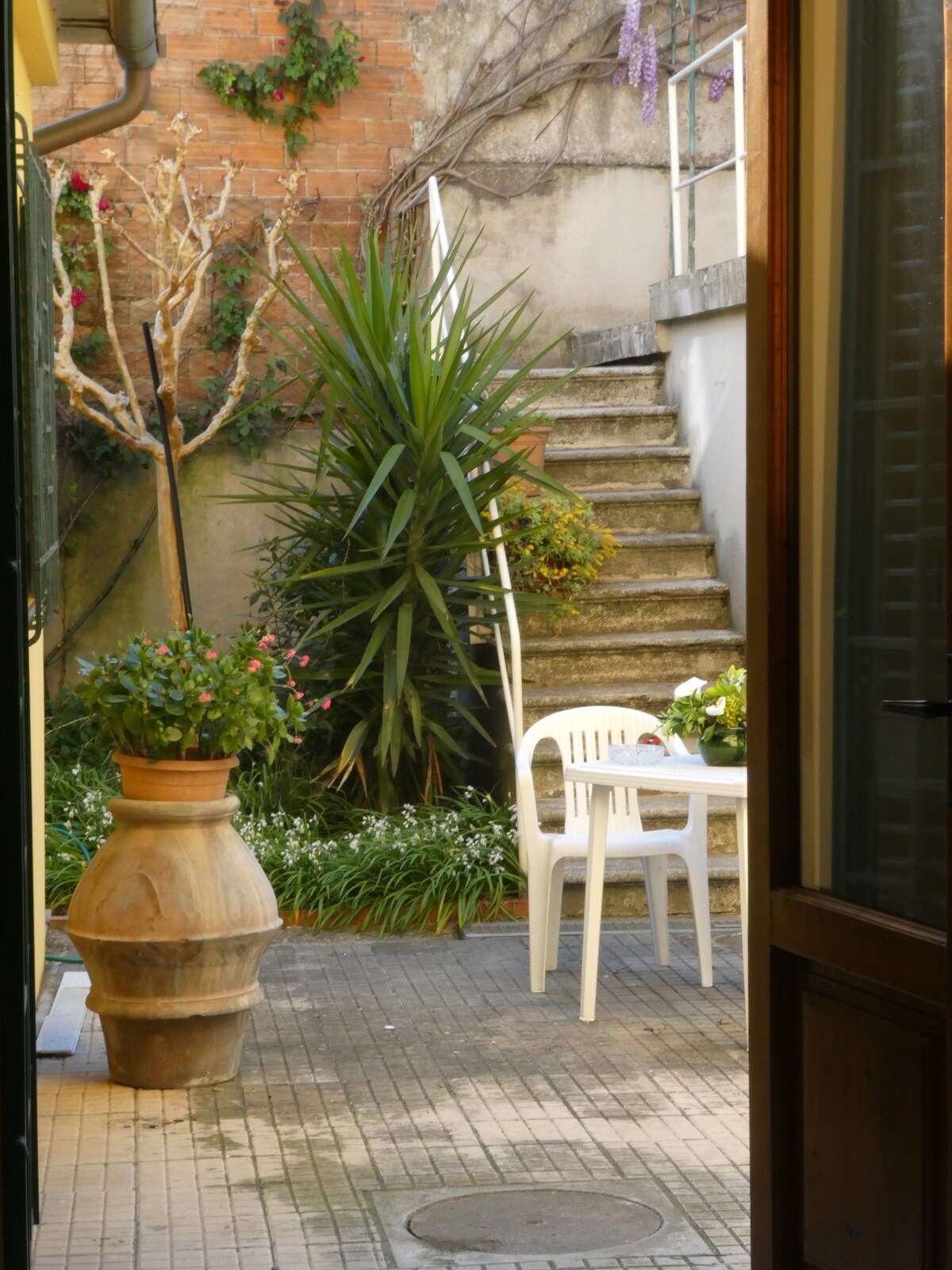 A tranquil garden area is visible, featuring a sturdy staircase leading to a higher level. A small table and chairs are set beside a large potted plant. Surrounded by greenery, the space reflects a serene atmosphere, ideal for outdoor relaxation.