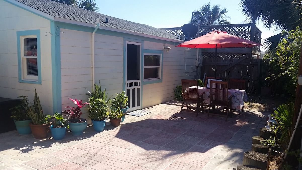 An outdoor patio area features a dining table surrounded by wooden chairs and shaded by a red umbrella. Colorful potted plants line the patio, complemented by a white cottage with blue trim and a fenced garden area in the background.