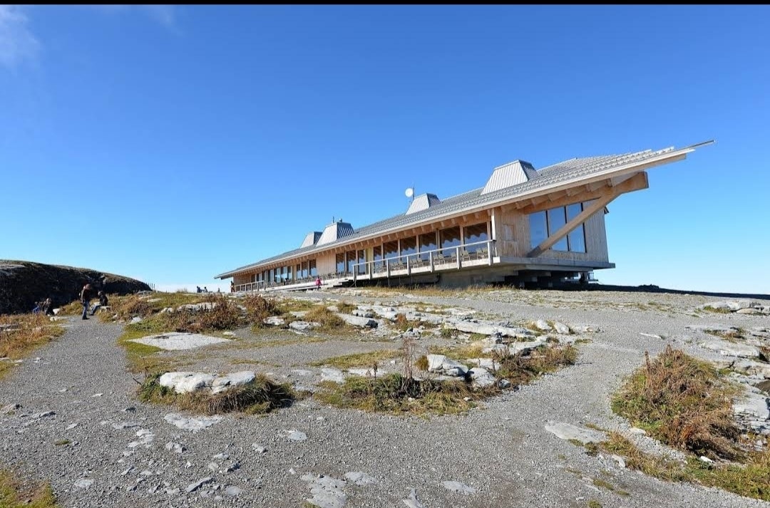 A modern wooden structure is perched on a rocky landscape, featuring a long design with multiple pitched roofs. Large windows line the front, providing expansive views. The surrounding area is marked by sparse vegetation and gravel pathways, highlighting the natural setting.