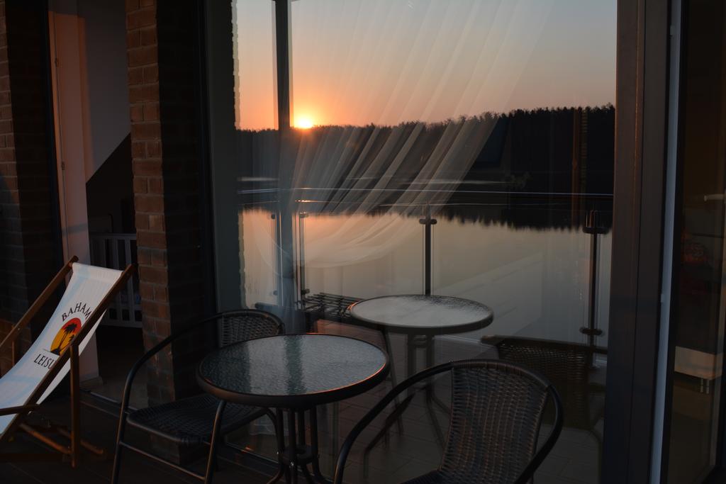 A view of the tranquil lake is framed by large glass doors, reflecting the warm colors of the sunset. Two wicker chairs and a round glass table are positioned on the balcony, enhancing the serene atmosphere of the outdoor space.