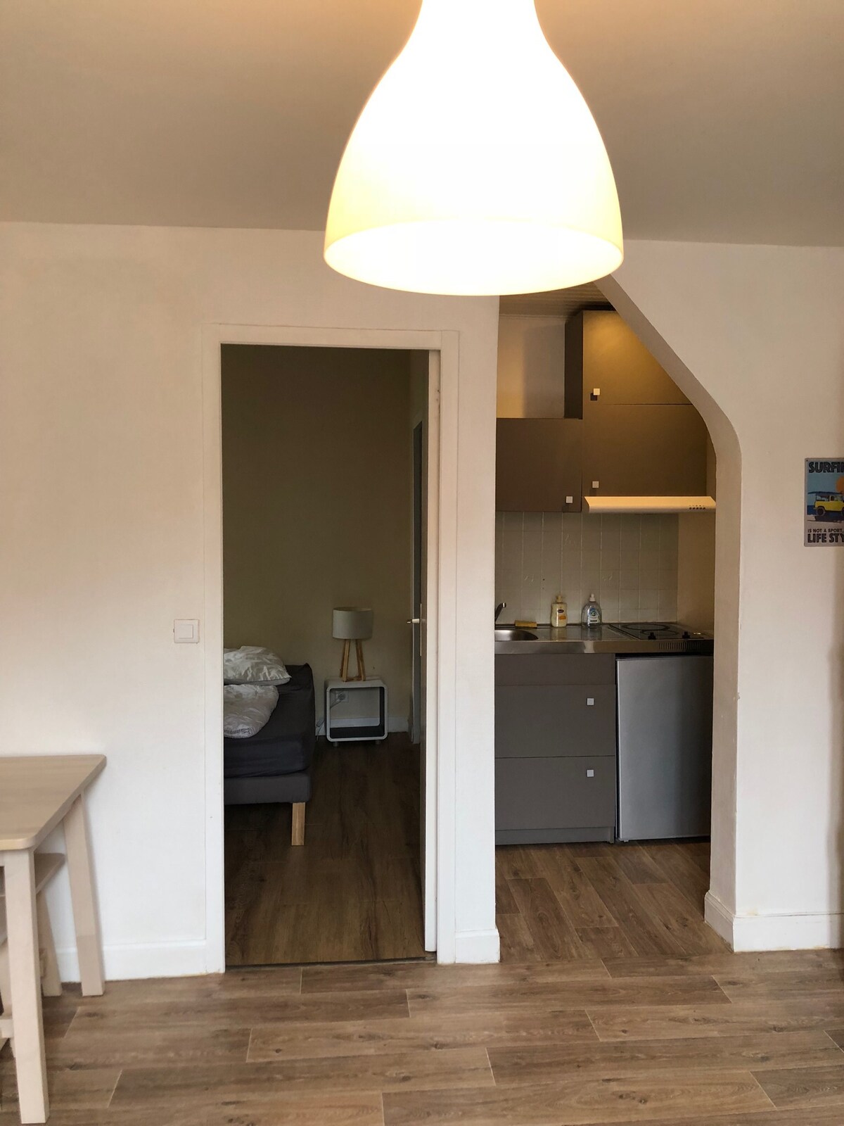 An open doorway leads to a cozy living area with a sofa and a small side table. The kitchen is partially visible, featuring modern cabinetry and essential appliances. Warm lighting from a stylish pendant illuminates the space, enhancing its inviting feel.
