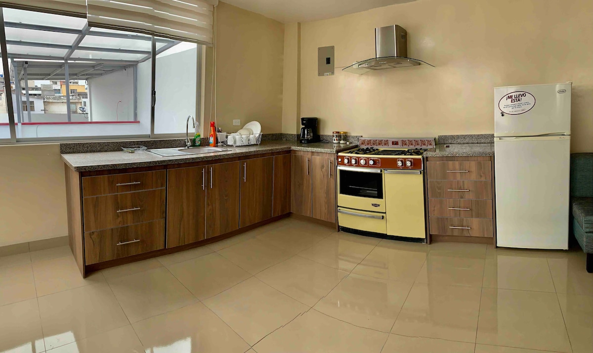A modern kitchen area is presented, featuring wooden cabinetry and a combination of a gas stove and an oven in a cream color. A white refrigerator stands nearby, while a countertop is equipped with a sink and kitchenware. Natural light enters through a large window.