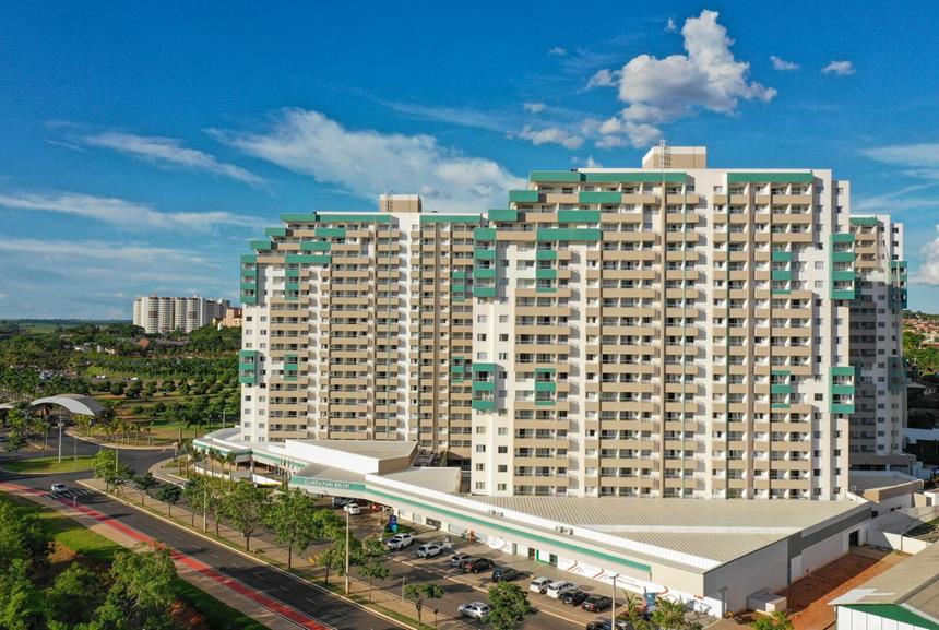 A modern multi-story building is depicted with a unique geometric facade featuring balconies. The structure is surrounded by landscaped greenery, providing a sense of space. A clear blue sky and scattered clouds create a pleasant backdrop, emphasizing the resort's welcoming environment.