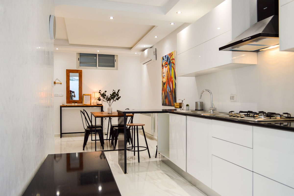 A modern kitchen and dining area feature sleek white cabinetry and a polished countertop. A wooden dining table is surrounded by black chairs. An art piece with vibrant colors adorns the wall, while natural light is provided by a nearby window.