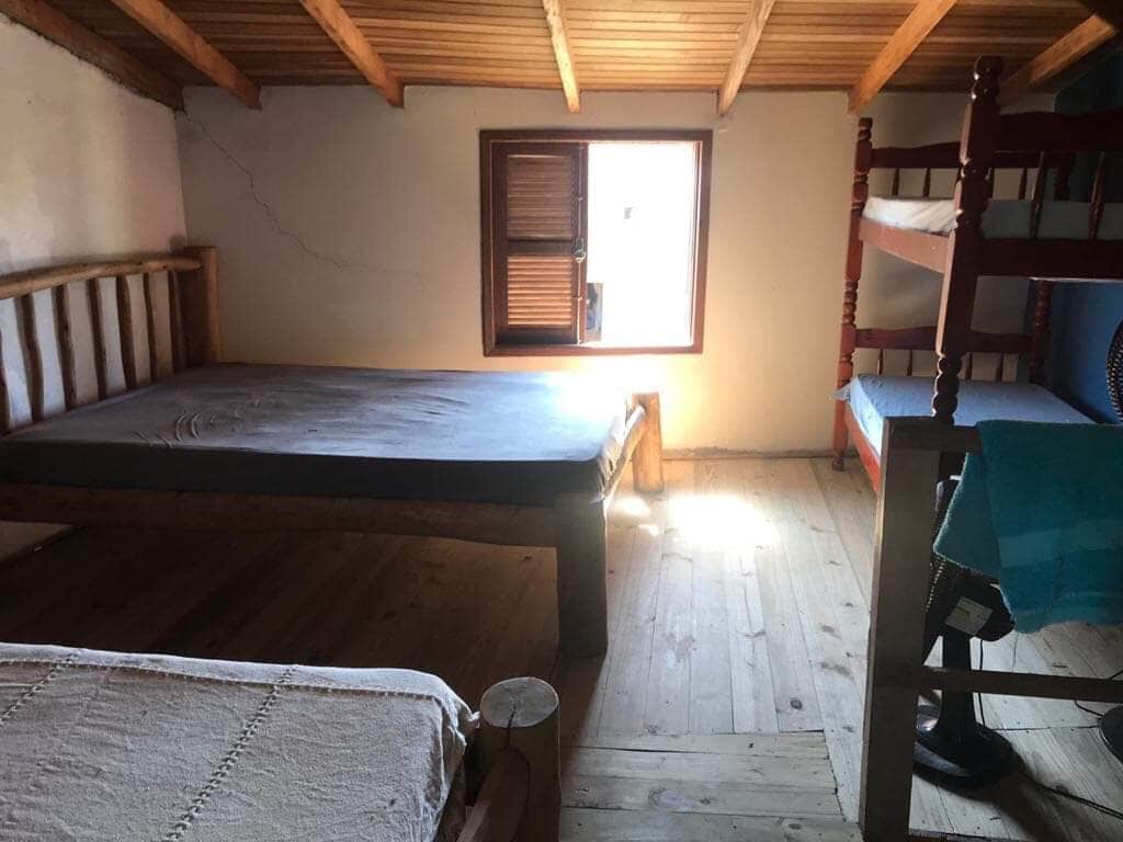 A simple sleeping area features a large bed and a bunk bed, both made from wood. Natural light enters through a small window, illuminating the wooden floor. A unique wooden stool is positioned near the bed, adding to the rustic charm of the space.