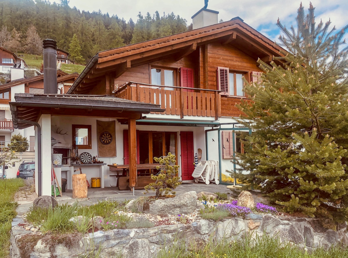 The chalet features a wooden exterior with a covered porch and balcony, surrounded by a landscaped garden. A stone pathway leads to a seating area, while a traditional chimney rises from the side. Lush greenery and colorful flowers enhance the inviting atmosphere.
